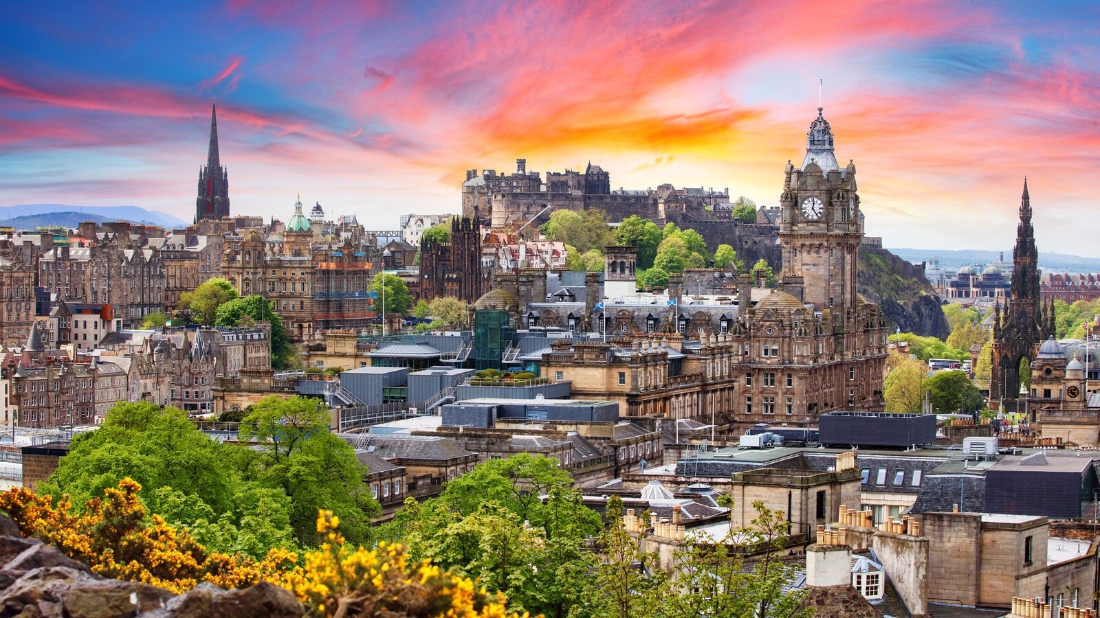 Edinburgh skyline at sunset with the Castle, Balmoral clock tower, and Scott Monument silhouetted against a vivid pink and orange sky